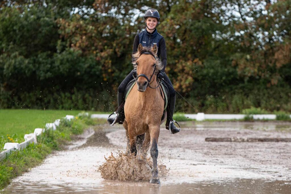Elev fra Ølgod Efterskole rider en brun hest gennem vand på udendørs ridebane.