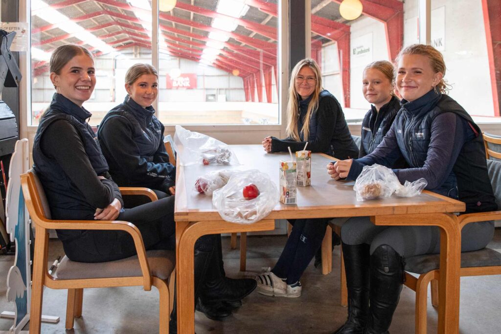 Elever fra Ølgod Efterskole sidder sammen ved et bord med ridehallen i baggrunden.