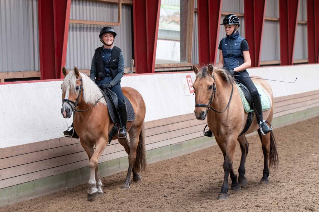 To elever fra Ølgod Efterskole rider side om side på heste i ridehallen.